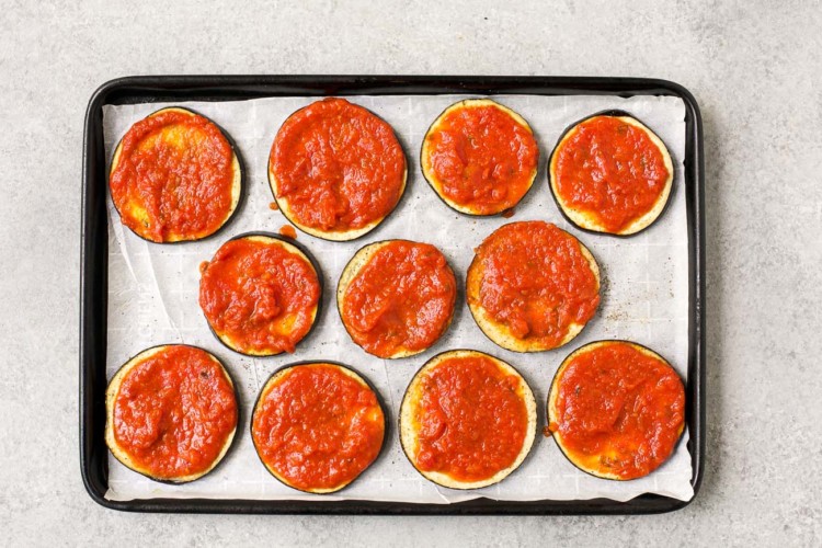 raw slices of eggplant with tomato sauce on the top on a baking sheet