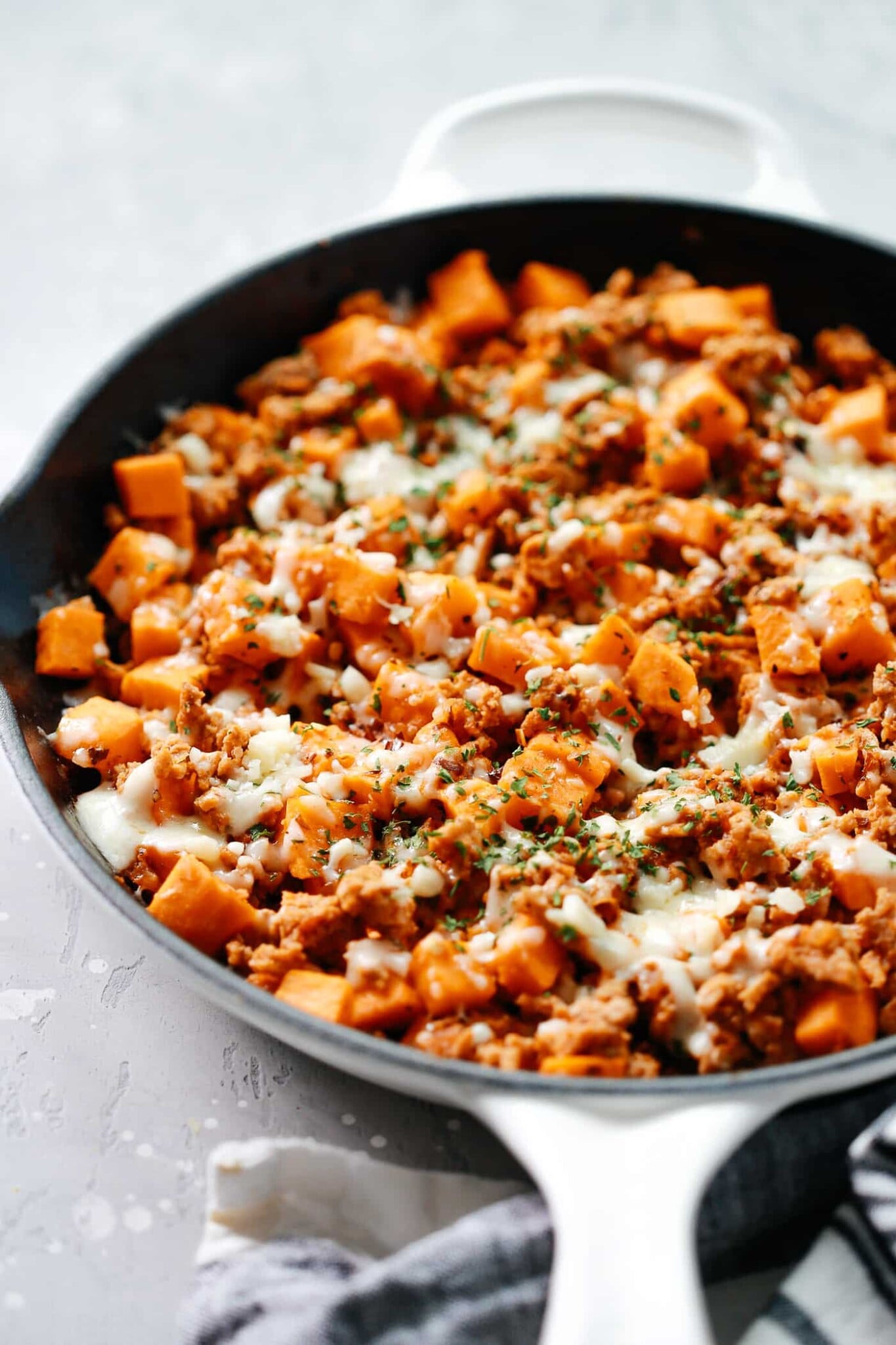 A Ground Turkey Sweet Potato Skillet filled with cooked ground meat, diced sweet potatoes, melted cheese, and chopped herbs sits on a light gray surface with a cloth partially underneath the pan.