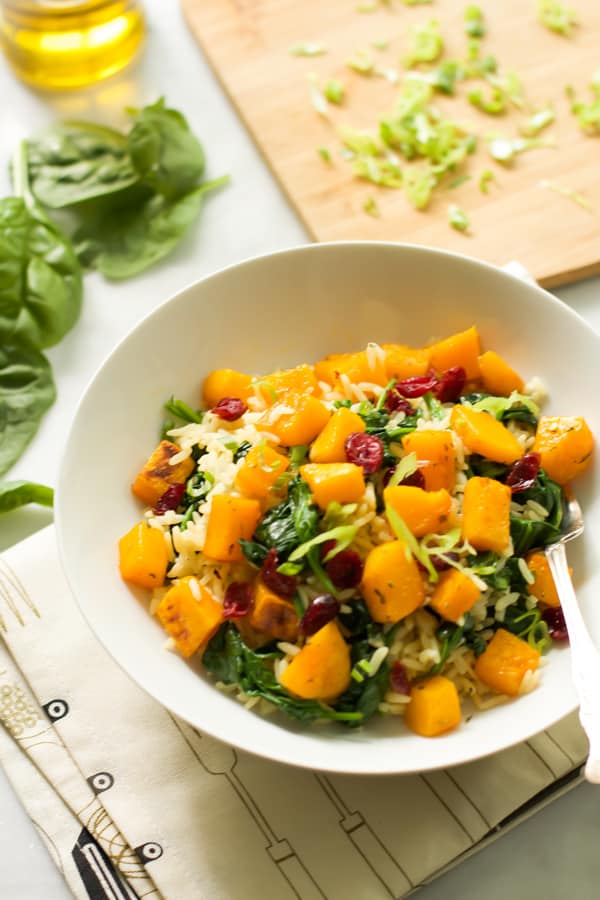 A bowl of rice with roasted butternut squash and dried cranberries with basil in the background.