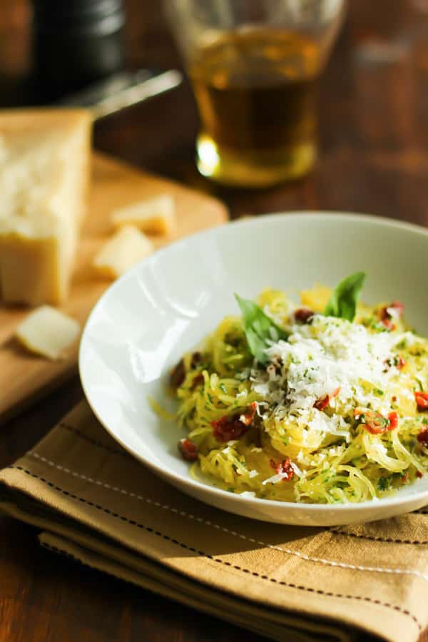 A bowl of spaghetti squash with sun-dried tomatoes and basil with parmesan in the background.