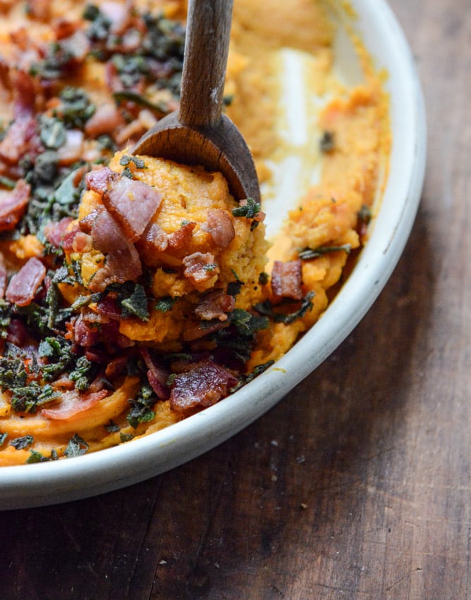 A close up of a wooden spoon scooping a bacon bourbon-whipped sweet potatoes with crispy sage in a casserole dish.