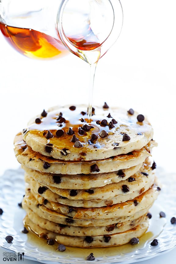 Guiltless Chocolate Chip Pancakes A stack of chocolate chip pancakes with syrup poured on top from a glass container.