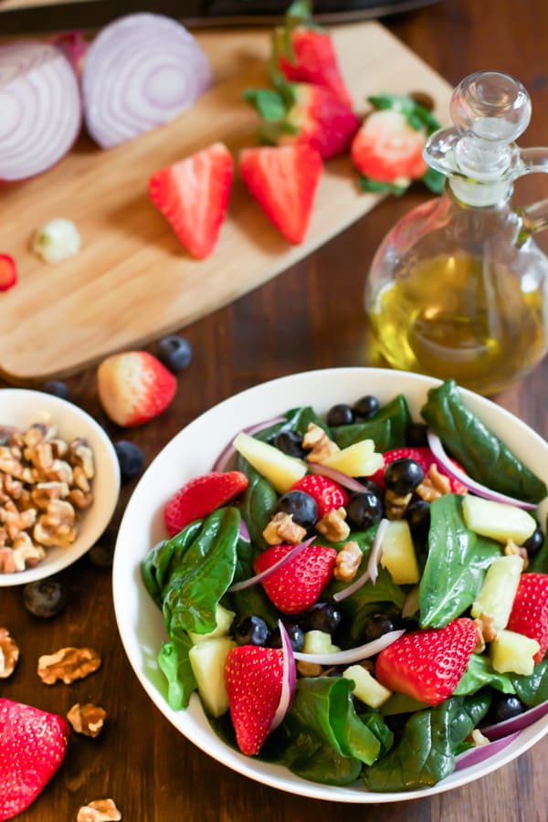 A bowl of spinach fruit salad with a cutting board with ingredients being prepped beside it.