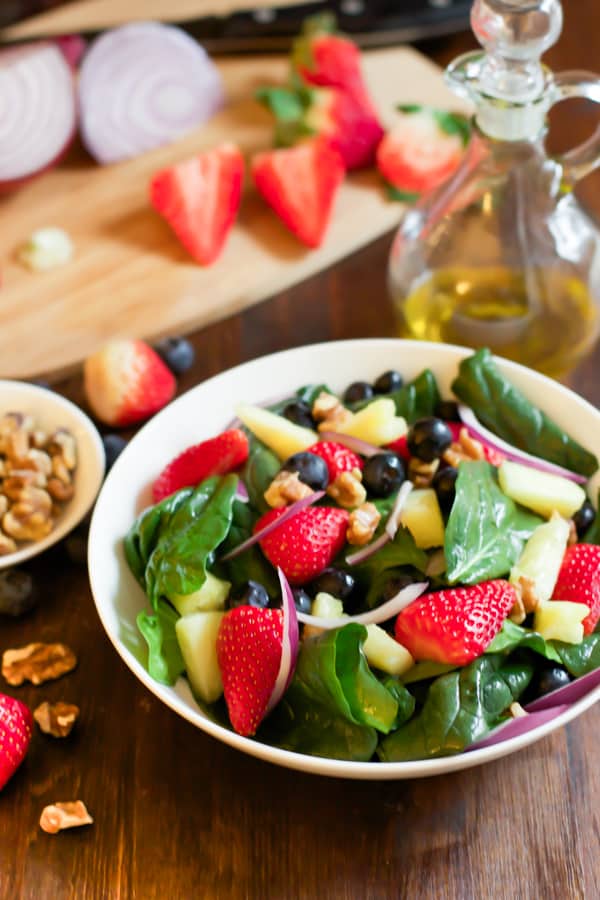 A bowl of spinach fruit salad with a cutting board with ingredients being prepped beside it.