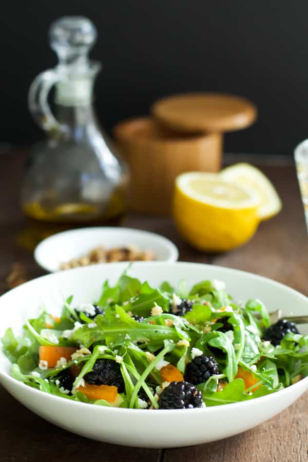 An arugula blackberry salad with oil, lemon, and salt in the background.