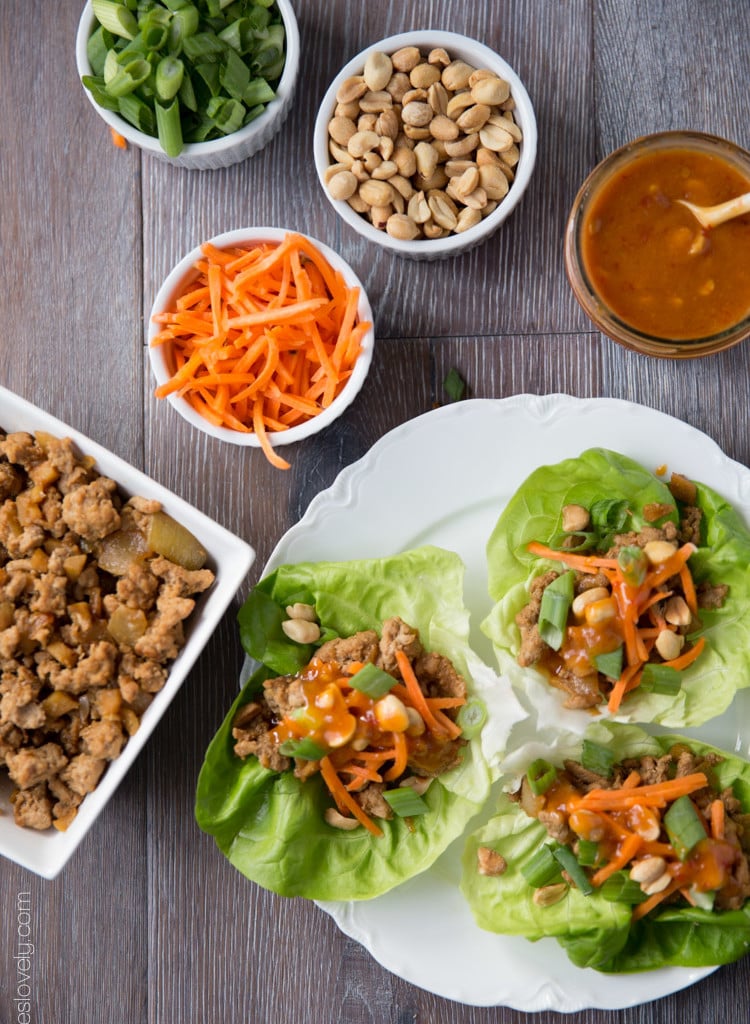 Three Asian turkey lettuce wrap on a plate with carrots, peanuts, green onions, sauce, and the filling in bowls beside it.