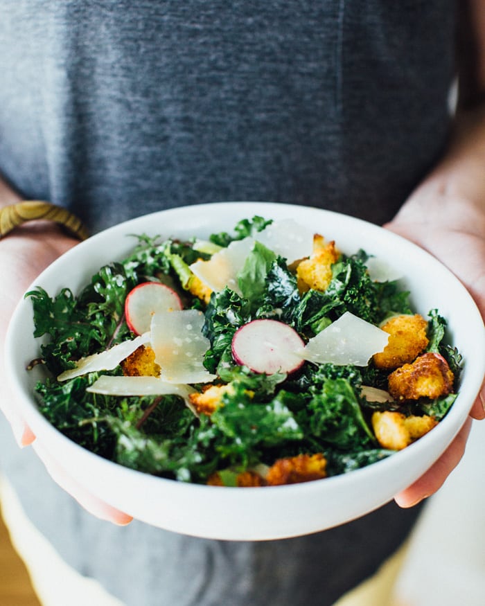 A set of hands holding a bowl of kale caesar salad with cornbread bits.