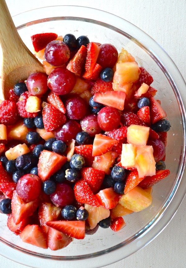 A glass bowl of secret fruit salad with a wooden spoon inside.