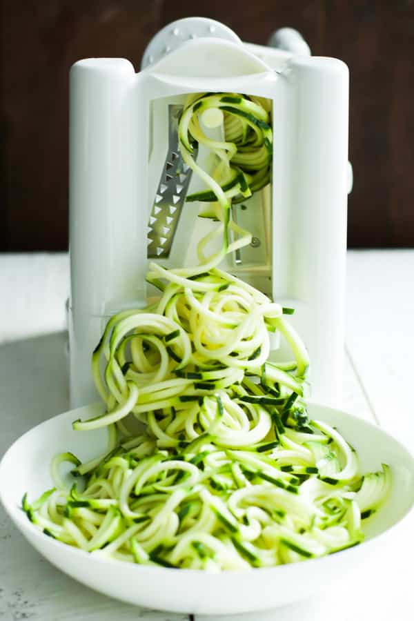 A vegetable spiralizer making zucchini noodles that are falling into a white bowl.