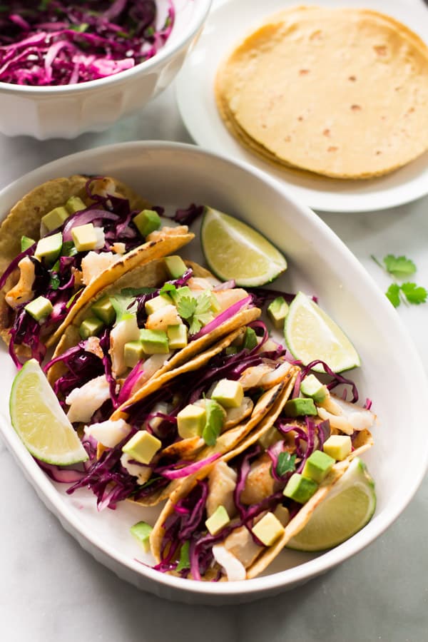 A serving bowl with four grilled fish tacos beside a plate of tortillas.