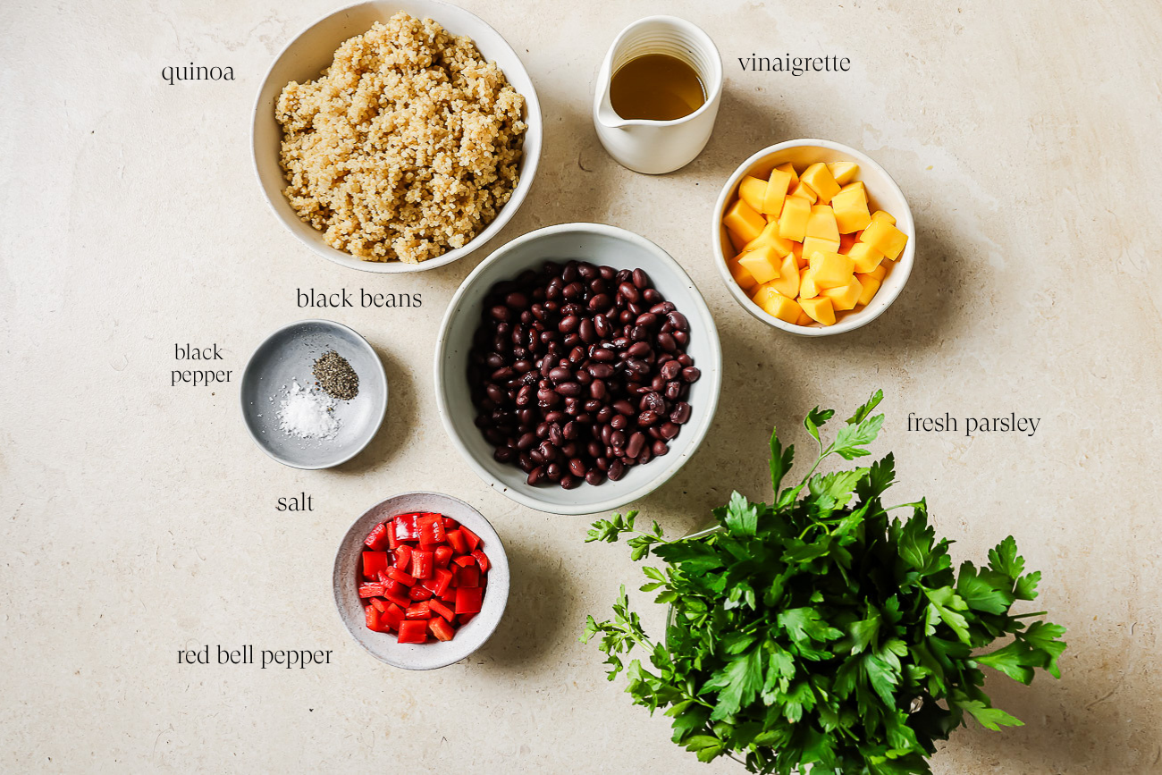 Bowls of mango quinoa salad with black beans, diced red bell pepper, salt, black pepper, a cup of vinaigrette, and a bunch of fresh parsley arranged on a light surface.