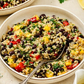 A vibrant mango quinoa salad featuring black beans, red bell peppers, yellow mango, and chopped parsley, served in a bowl with a spoon and a lemon wedge on the side.