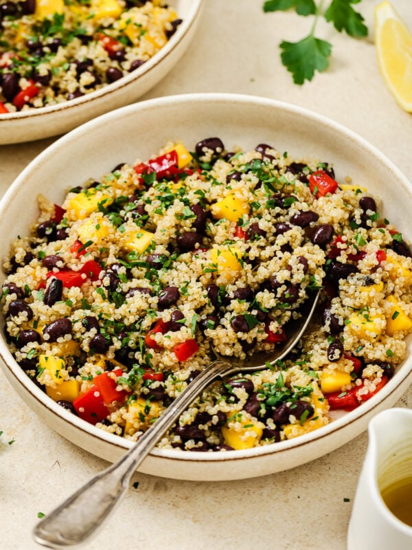 A vibrant mango quinoa salad featuring black beans, red bell peppers, yellow mango, and chopped parsley, served in a bowl with a spoon and a lemon wedge on the side.