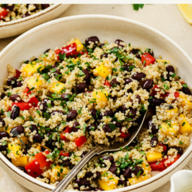 A bowl of mango quinoa salad with black beans, red bell peppers, chopped herbs, and a fork. Text above reads: Mango Quinoa Salad - Easy, fresh, and ready in just 10 minutes to prep this delicious mango quinoa salad.
