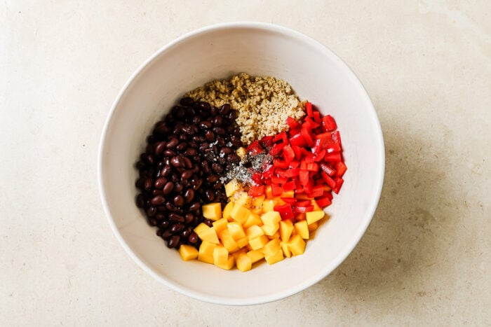 A white bowl contains separate portions of black beans, cooked quinoa, diced red bell pepper, diced mango, and seasoning on a light countertop—perfect for assembling a fresh mango quinoa salad.