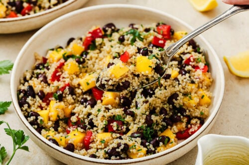 A close-up of a vibrant mango quinoa salad with black beans, diced mango, red bell peppers, and herbs. A spoon lifts a portion from the bowl, while lemon wedges and fresh parsley sit nearby.