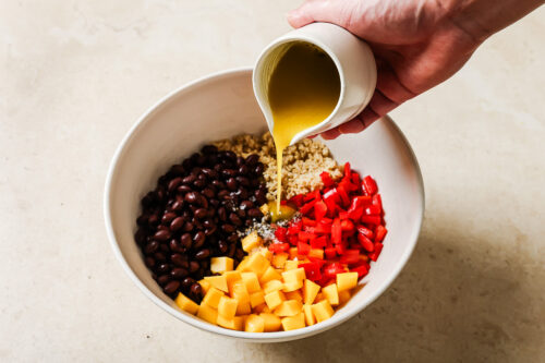 A hand pours yellow dressing from a small white jug into a vibrant mango quinoa salad, filled with black beans, chopped red bell pepper, and diced mango on a light countertop.