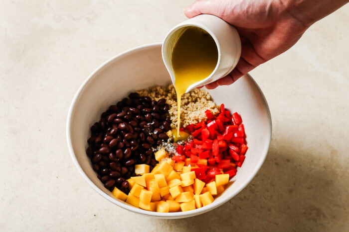 A hand pours yellow dressing from a small white jug into a vibrant mango quinoa salad, filled with black beans, chopped red bell pepper, and diced mango on a light countertop.