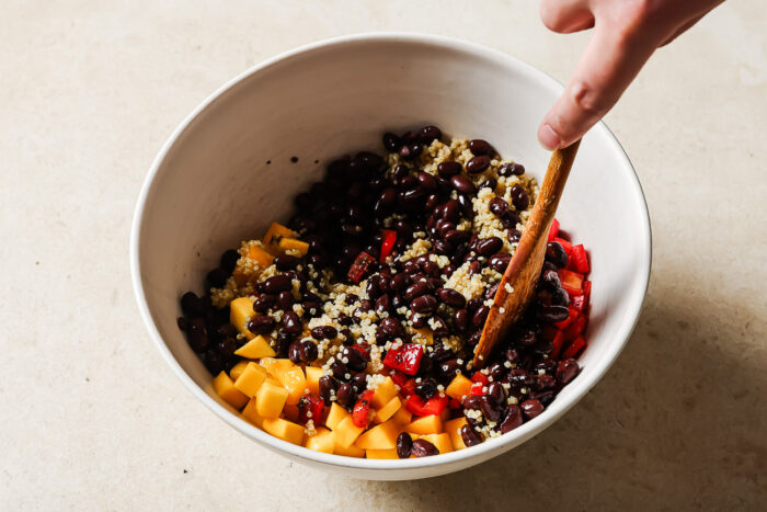 A hand stirs a bowl filled with black beans, quinoa, diced yellow mango, and red bell peppers—classic ingredients for a vibrant mango quinoa salad—using a wooden spoon on a light countertop.