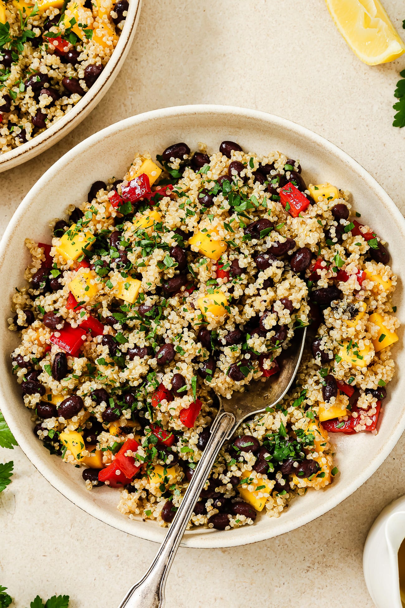 A bowl of mango quinoa salad with black beans, diced red and yellow bell peppers, chopped herbs, and a spoon, placed on a light surface with lemon wedges and parsley nearby.