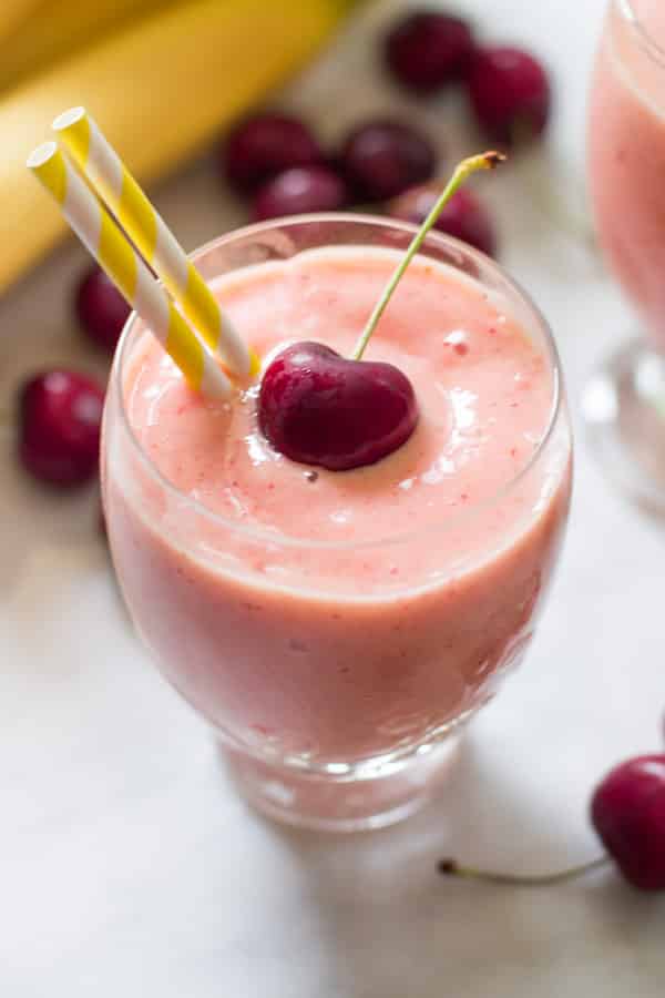Close up photo of a glass of cherry pineapple smoothie with two straws inserted and a cherry on top as garnish. 