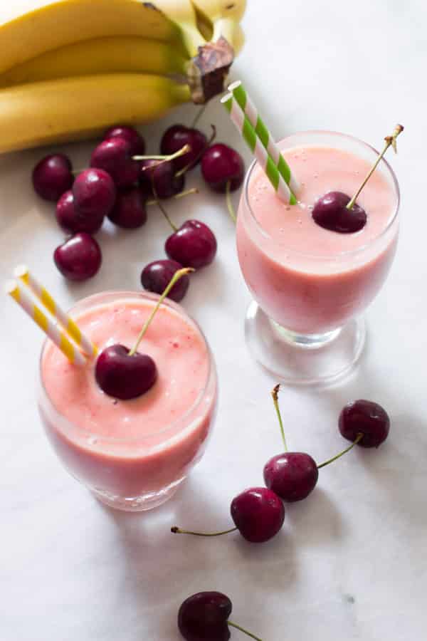 Two glasses of cherry pineapple smoothies beside cherries and bananas.
