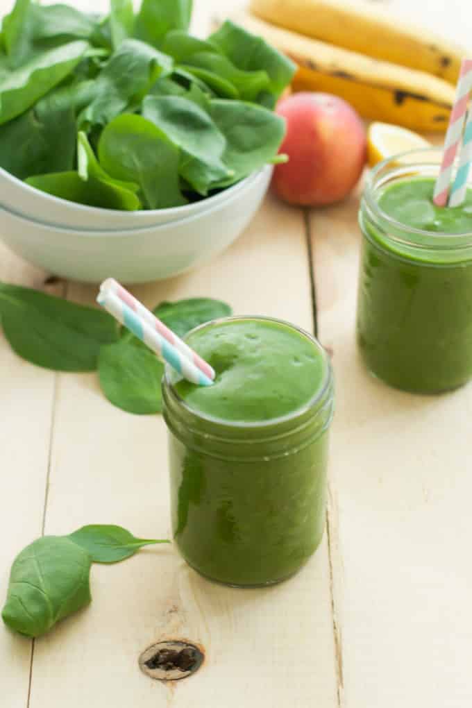 Two glasses of peach green smoothie beside an apple, bananas, and a bowl of spinach.