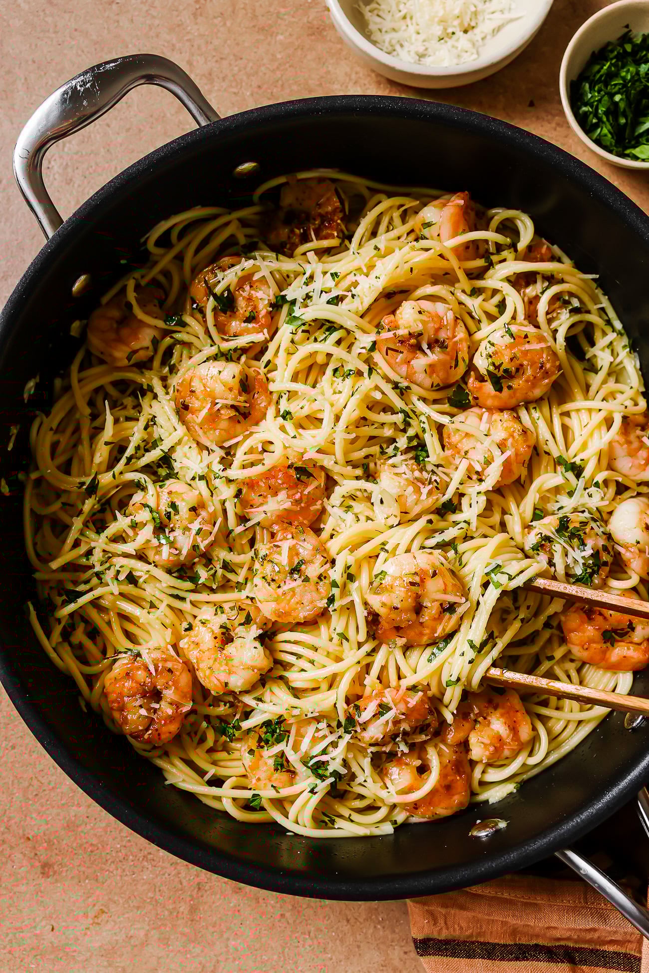 A skillet filled with Garlic Shrimp Spaghetti, saut&eacute;ed shrimp, chopped parsley, and grated cheese, with tongs resting on top. Small bowls of cheese and herbs are nearby on a light brown surface.