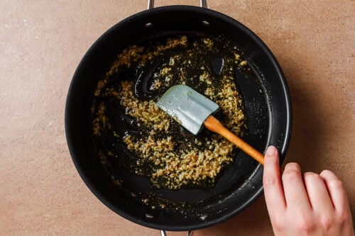 A hand stirs minced garlic and herbs in oil with a spatula in a black skillet on a brown countertop, prepping the flavors for Garlic Shrimp Spaghetti.