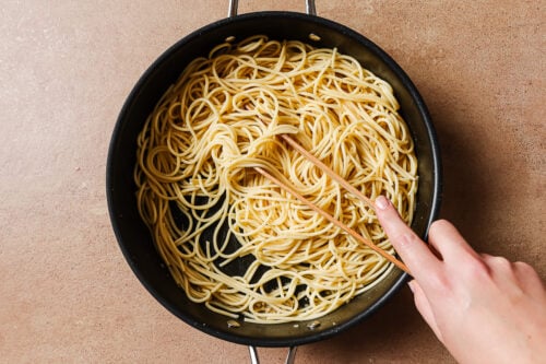 A hand uses chopsticks to stir cooked garlic shrimp spaghetti noodles in a black pan on a brown countertop.
