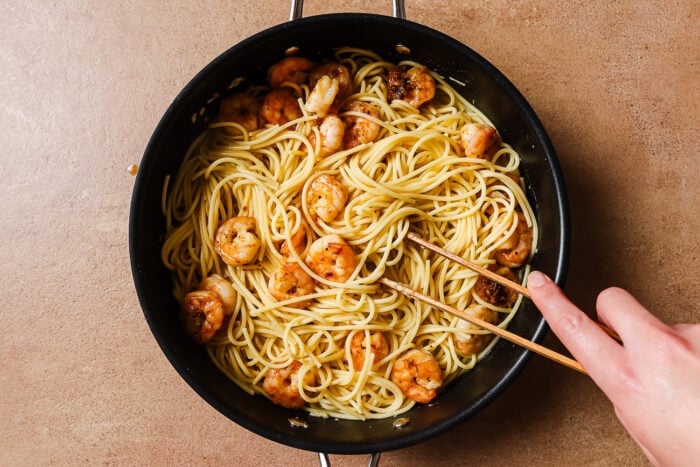 A hand holding chopsticks stirs Garlic Shrimp Spaghetti in a black pan on a brown countertop.