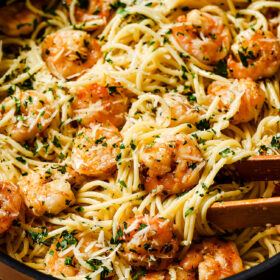A skillet filled with Garlic Shrimp Spaghetti, sautéed shrimp, chopped parsley, and grated parmesan cheese, with wooden tongs mixing the flavorful ingredients.