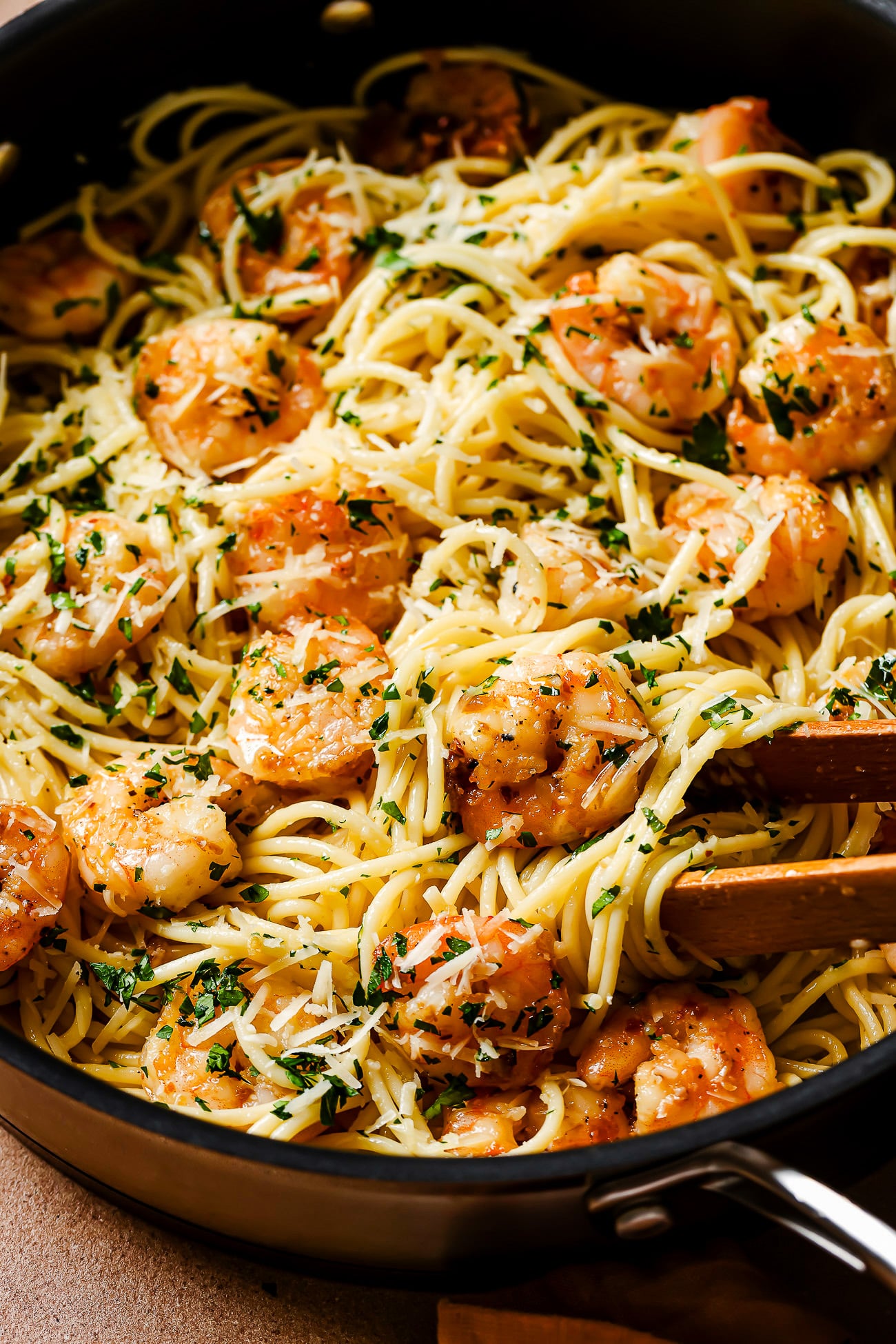 A skillet filled with Garlic Shrimp Spaghetti, saut&eacute;ed shrimp, chopped parsley, and grated parmesan cheese, with wooden tongs mixing the flavorful ingredients.