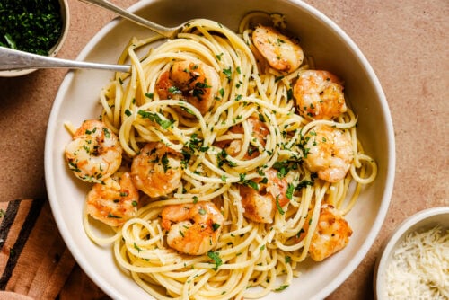 A bowl of Garlic Shrimp Spaghetti topped with chopped parsley, with a fork and spoon nestled inside. A small bowl of shredded cheese sits in the background, ready for sprinkling.