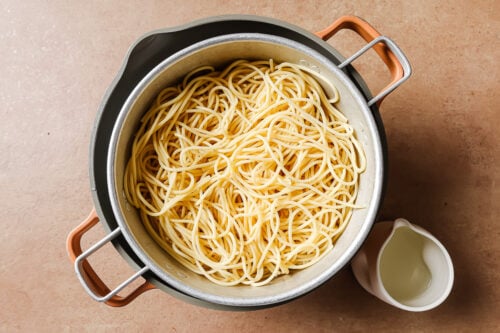 A pot filled with cooked Garlic Shrimp Spaghetti sits on a beige surface. Next to it is a small white pitcher containing pasta water.
