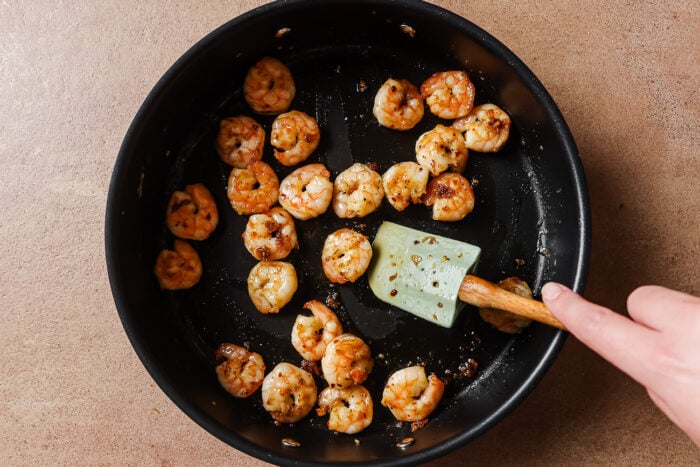 A hand uses a spatula to cook seasoned shrimp for Garlic Shrimp Spaghetti in a black frying pan on a brown countertop.