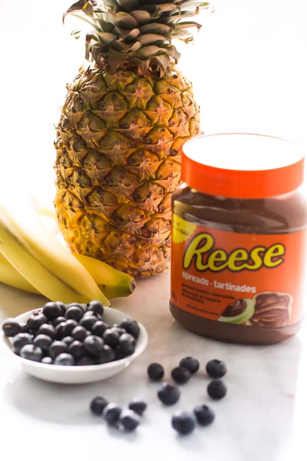 A plate of blueberries, two bananas, a pineapple, and a jar of reese spread.