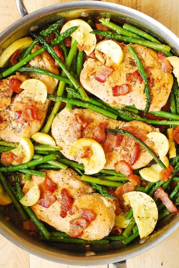 An overhead view of chicken, asparagus, and squash topped with bacon in a skillet.