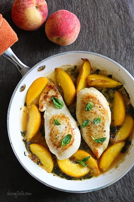 An overhead view of two chicken breasts and peach slices in a white skillet.