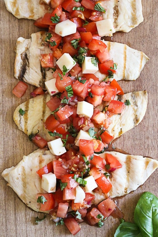 Four grilled chicken cutlets topped with tomatoes, basil, and mozzarella cheese on a wooden cutting board.