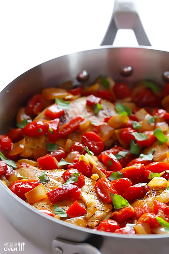 A closeup of Italian chicken topped with tomatoes, onions, and fresh basil inside a metal skillet.