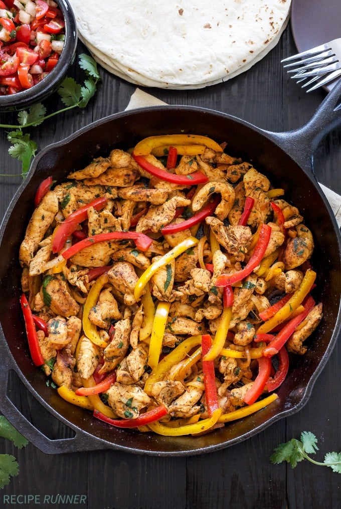 Overhead view of chicken and bell peppers in a cast iron skillet with tortillas and Pico de Gallo on the side.