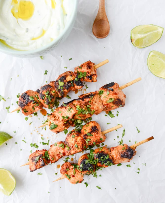 Four chili garlic chicken skewers on parchment paper with a bowl of Greek yogurt dip on the side.
