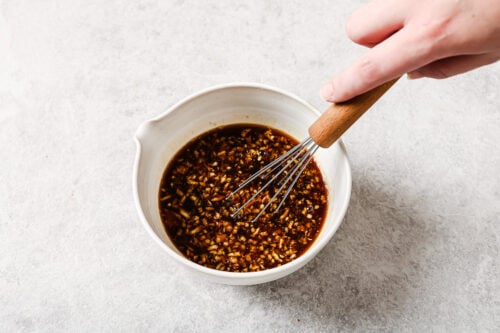 A hand holding a whisk stirs a dark, chunky Grilled Pineapple Chicken sauce in a white bowl on a light, textured surface.