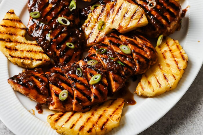 Grilled Pineapple Chicken breasts glazed with a dark sauce, garnished with sliced green onions, and served alongside grilled pineapple slices on a white plate.