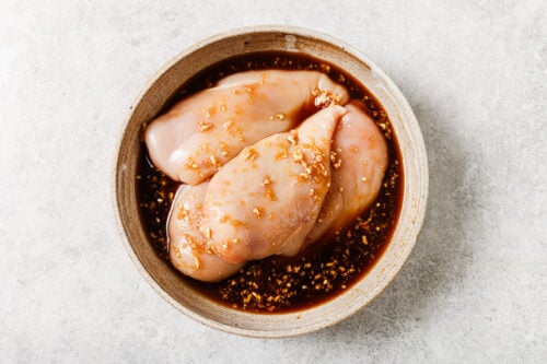 Raw chicken breasts marinating in a light brown sauce with minced garlic, seasonings, and hints of sweetness for Grilled Pineapple Chicken, placed in a round beige bowl on a light stone surface.