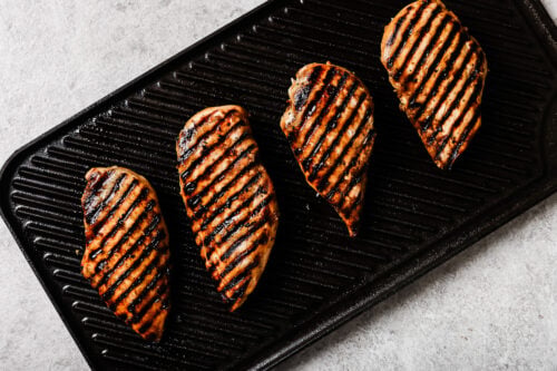 Four Grilled Pineapple Chicken breasts with visible char marks are arranged in a row on a ridged grill pan against a light background.