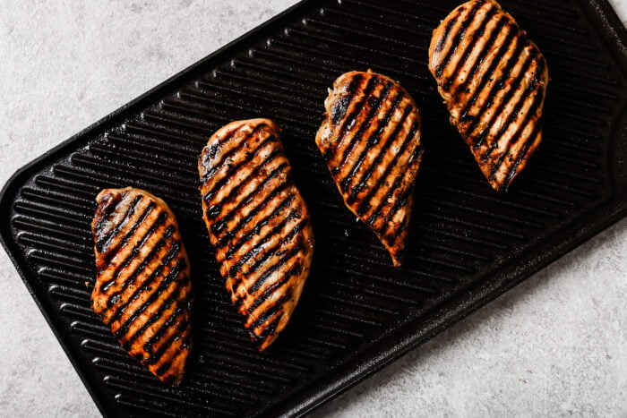Four Grilled Pineapple Chicken breasts with visible char marks are arranged in a row on a ridged grill pan against a light background.