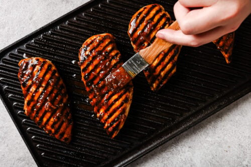 A hand brushes barbecue sauce onto Grilled Pineapple Chicken breasts with grill marks, arranged in a row on a black grill pan.