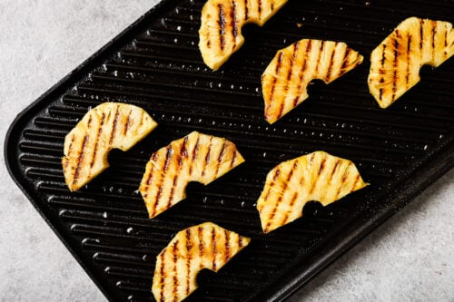 Grilled pineapple slices with char marks are arranged on a black grill pan, set on a light gray surface—perfect as a sweet side for your favorite Grilled Pineapple Chicken.